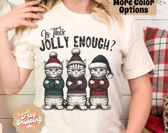 May include: A white t-shirt with the text "Is This Jolly Enough?" and a graphic of three cats wearing Santa hats and sweaters. The cats are standing in a row and have their arms crossed.
