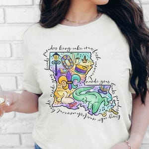 Op de afbeelding: Een wit T-shirt met een kleurrijke illustratie van de staat Louisiana. De illustratie bevat Mardi Gras-parels, een koningentaart, een rivierkreeft, een fleur-de-lis en andere symbolen van de Louisiana-cultuur. De tekst "beads parades king cake crawfish carnival mardi gras beads parades king cake crawfish carnival mardi gras" omringt de illustratie.