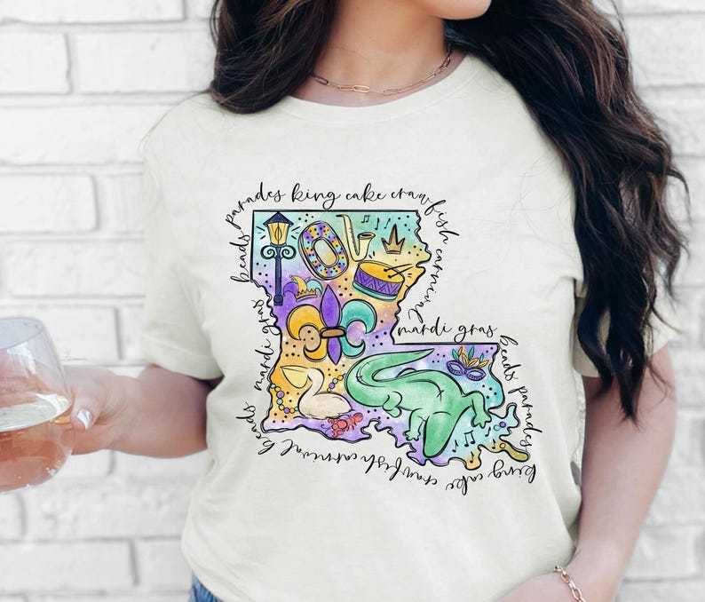 Op de afbeelding: Een wit T-shirt met een kleurrijke illustratie van de staat Louisiana. De illustratie bevat Mardi Gras-parels, een koningentaart, een rivierkreeft, een fleur-de-lis en andere symbolen van de Louisiana-cultuur. De tekst "beads parades king cake crawfish carnival mardi gras beads parades king cake crawfish carnival mardi gras" omringt de illustratie.