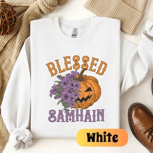 以下が含まれることがあります： 白いスウェットシャツに、紫色の文字で「Blessed Samhain」と書いてあり、紫色の花がついたジャック・オー・ランタンが描かれています。