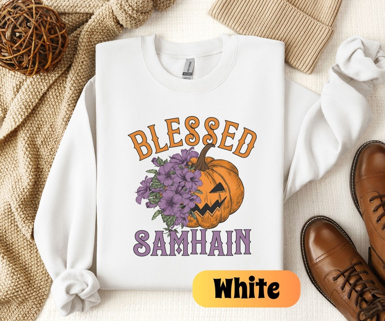以下が含まれることがあります： 白いスウェットシャツに、紫色の文字で「Blessed Samhain」と書いてあり、紫色の花がついたジャック・オー・ランタンが描かれています。