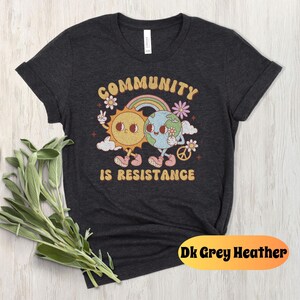 May include: Dark grey heather t-shirt with a graphic of a sun and earth holding hands. The graphic is surrounded by flowers and the text "Community is Resistance".
