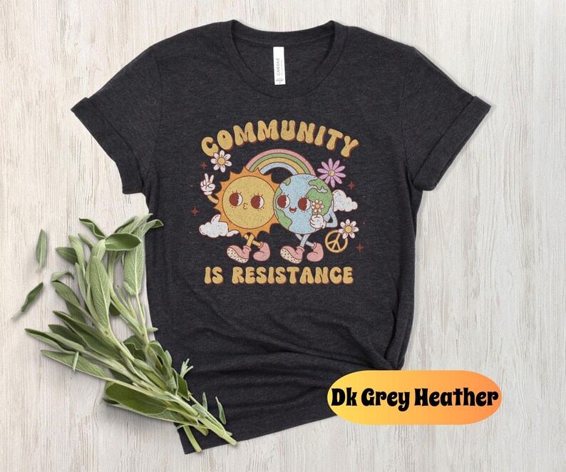 May include: Dark grey heather t-shirt with a graphic of a sun and earth holding hands. The graphic is surrounded by flowers and the text "Community is Resistance".