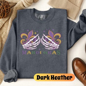 May include: Dark heather grey sweatshirt with a graphic of two skeletal hands holding purple and gold fleur-de-lis. The text "Mardi Gras" is printed below the hands.