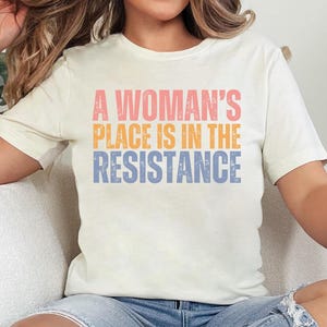 May include: Cream-colored t-shirt with the text "A Woman's Place Is In The Resistance" in pink, orange, and blue. The shirt has short sleeves and a crew neck. The shirt is made of a soft material.