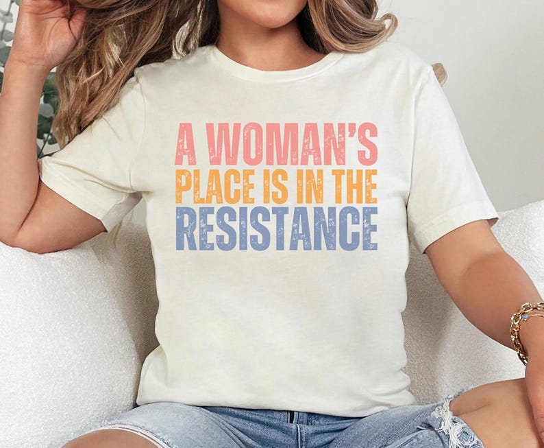 May include: Cream-colored t-shirt with the text "A Woman's Place Is In The Resistance" in pink, orange, and blue. The shirt has short sleeves and a crew neck. The shirt is made of a soft material.