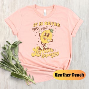 May include: A pink t-shirt with a retro-style graphic of a smiling lemon with the text "It is never easy peasy lemon squeezy".