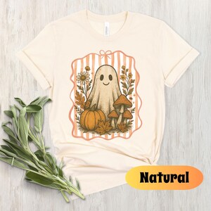 May include: Cream-colored t-shirt featuring a whimsical ghost illustration with a pumpkin, mushrooms, and fall foliage. The design is framed by a peach-colored striped border. The word "Natural" is displayed in a yellow and orange oval.