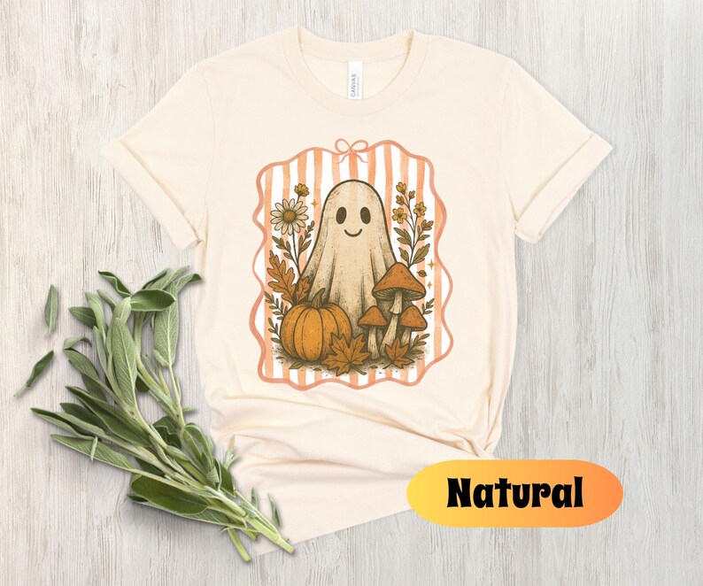 May include: Cream-colored t-shirt featuring a whimsical ghost illustration with a pumpkin, mushrooms, and fall foliage. The design is framed by a peach-colored striped border. The word "Natural" is displayed in a yellow and orange oval.