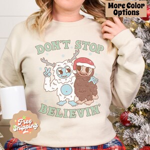 May include: A cream-colored sweatshirt with a Christmas-themed graphic. The graphic features two cartoon characters, a yeti and a sasquatch, with the text "Don't Stop Believin'" below them.