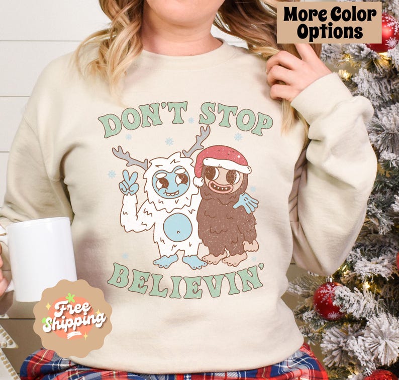May include: A cream-colored sweatshirt with a Christmas-themed graphic. The graphic features two cartoon characters, a yeti and a sasquatch, with the text "Don't Stop Believin'" below them.
