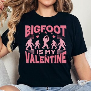 May include: Black t-shirt with a pink graphic that says "BIGFOOT IS MY VALENTINE". The graphic features a line of Bigfoot figures with hearts and stars.