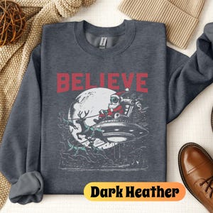 May include: Dark heather grey sweatshirt with a graphic of Santa Claus flying in a UFO with reindeer. The text "BELIEVE" is printed in red above the graphic.