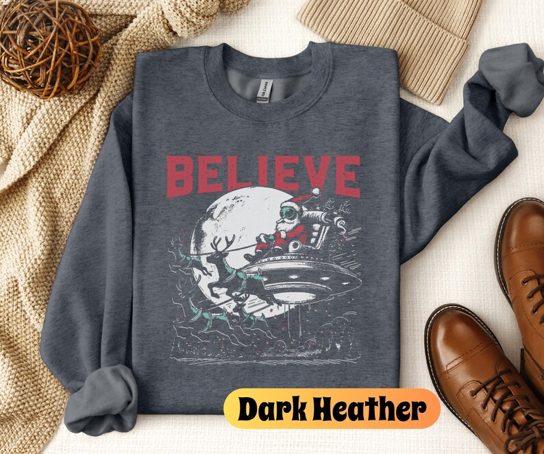 May include: Dark heather grey sweatshirt with a graphic of Santa Claus flying in a UFO with reindeer. The text "BELIEVE" is printed in red above the graphic.