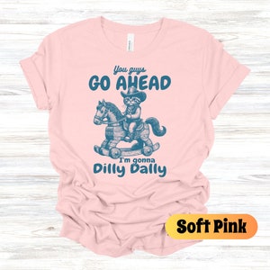 May include: A soft pink t-shirt with a blue graphic of a raccoon wearing a cowboy hat and riding a rocking horse. The text on the shirt reads "You guys go ahead I'm gonna dilly dally".