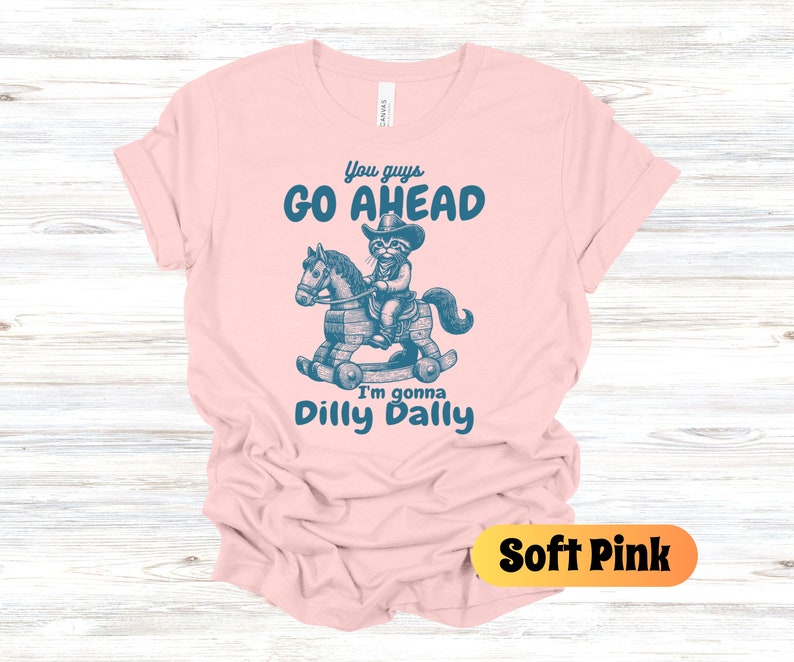 May include: A soft pink t-shirt with a blue graphic of a raccoon wearing a cowboy hat and riding a rocking horse. The text on the shirt reads "You guys go ahead I'm gonna dilly dally".