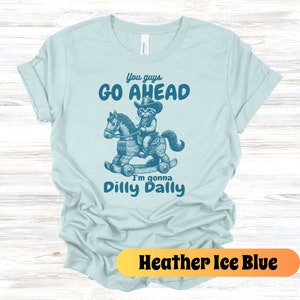 May include: A light blue t-shirt with a graphic of a cat riding a rocking horse. The text on the shirt reads "You guys go ahead I'm gonna dilly dally".