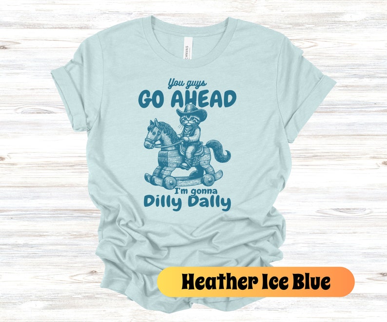 May include: A light blue t-shirt with a graphic of a cat riding a rocking horse. The text on the shirt reads "You guys go ahead I'm gonna dilly dally".