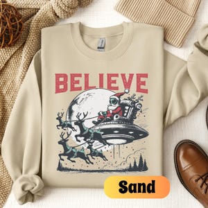 May include: A beige sweatshirt with the text "BELIEVE" in red letters. A cartoon image of Santa Claus wearing a space helmet and riding a flying saucer pulled by reindeer is below the text. The word "Sand" is written in yellow on a white background in the lower right corner of the image.