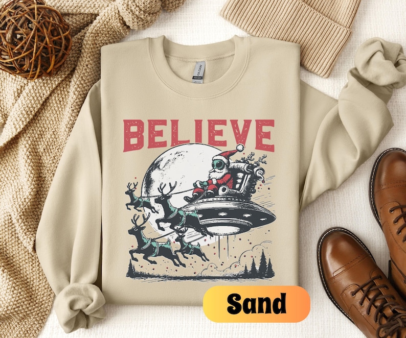 May include: A beige sweatshirt with the text "BELIEVE" in red letters. A cartoon image of Santa Claus wearing a space helmet and riding a flying saucer pulled by reindeer is below the text. The word "Sand" is written in yellow on a white background in the lower right corner of the image.