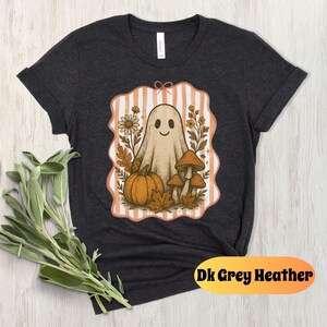 May include: Dark grey heather t-shirt featuring a whimsical ghost design. The graphic includes a smiling ghost, pumpkin, mushrooms, and fall foliage within a striped frame. The text "Dk Grey Heather" is at the bottom.