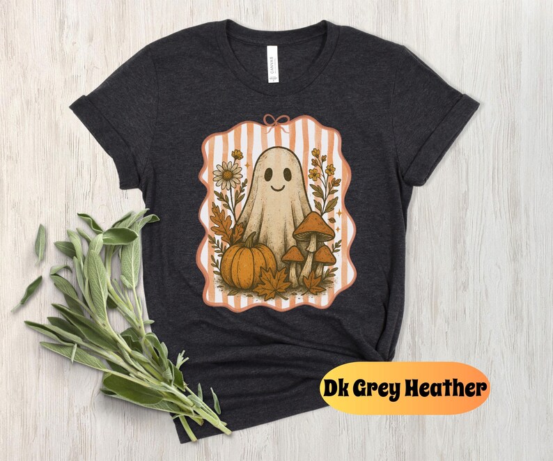 May include: Dark grey heather t-shirt featuring a whimsical ghost design. The graphic includes a smiling ghost, pumpkin, mushrooms, and fall foliage within a striped frame. The text "Dk Grey Heather" is at the bottom.