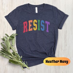 May include: A heather navy blue t-shirt with the word "RESIST" printed in rainbow colors across the front.