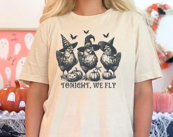 May include: A beige t-shirt with a graphic of three chickens wearing witch hats and holding pumpkins. The text "Tonight, We Fly" is printed below the chickens.