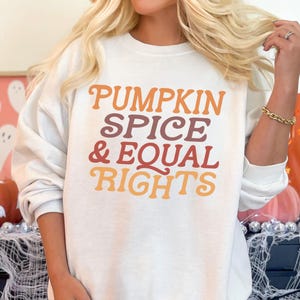 May include: White sweatshirt with the text "PUMPKIN SPICE & EQUAL RIGHTS" in orange, purple, and brown lettering. The sweatshirt has long sleeves and a crew neck. The image has a fall theme with pumpkins and ghosts in the background.