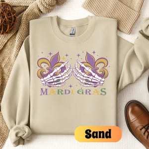 May include: Sand-colored sweatshirt with a purple, green, and gold Mardi Gras design featuring two skeletal hands holding fleur-de-lis. The text "Mardi Gras" is printed below the design.