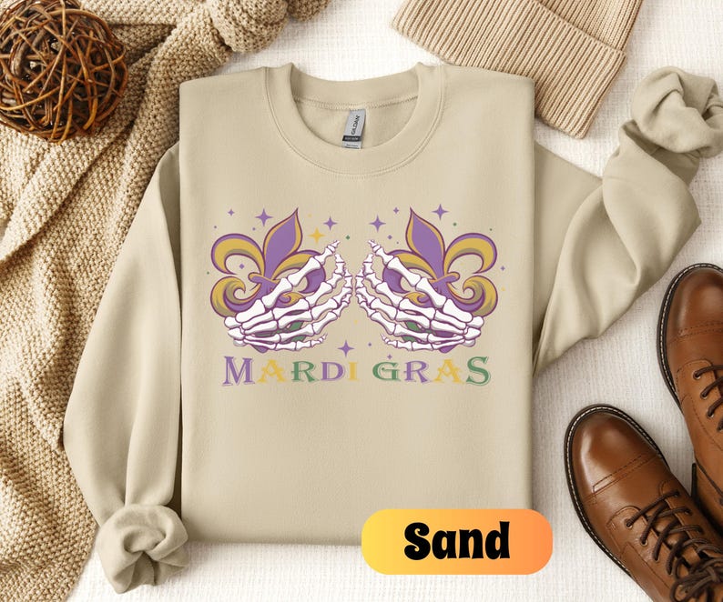 May include: Sand-colored sweatshirt with a purple, green, and gold Mardi Gras design featuring two skeletal hands holding fleur-de-lis. The text "Mardi Gras" is printed below the design.