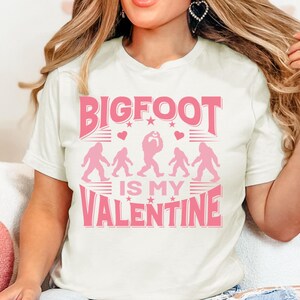 May include: White t-shirt with a pink graphic that says "BIGFOOT IS MY VALENTINE". The graphic features a line of Bigfoot figures with hearts.