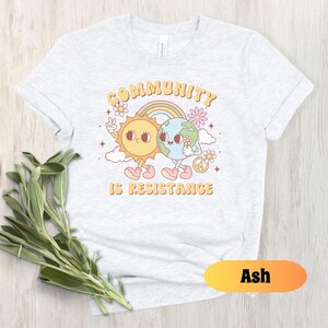 May include: A light gray t-shirt with a graphic of a sun and earth holding hands. The text "Community is Resistance" is printed below the graphic. The shirt is available in the color Ash.