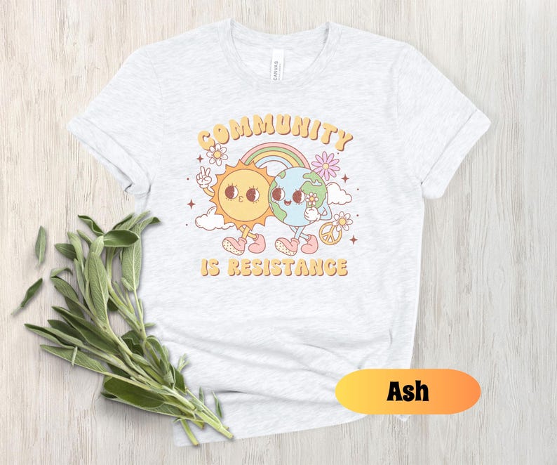 May include: A light gray t-shirt with a graphic of a sun and earth holding hands. The text "Community is Resistance" is printed below the graphic. The shirt is available in the color Ash.