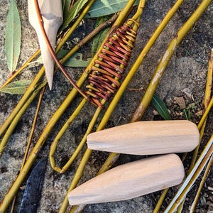 Può includere: Primo piano di strumenti e materiali per la tessitura di vimini. Gli strumenti includono due pioli di legno e un mazzuolo di legno. I materiali includono rami di salice e un pezzo di salice intrecciato.