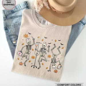 Spooky Halloween Nurse Skeleton shirt featuring dancing skeletons wearing nurse hats with medical supplies, IV drip, pumpkins, and fall leaves on an ivory tee.
