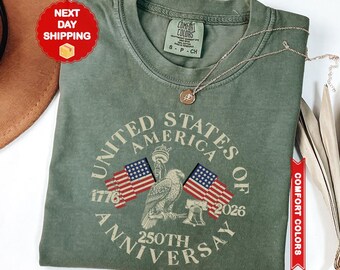 以下が含まれることがあります： モスグリーンのクルーネックTシャツ。United States of America 250th Anniversary、1776、2026の文字がプリントされています。デザインには2つのアメリカ国旗、ハクトウワシ、自由の女神像、リバティベルが含まれています。