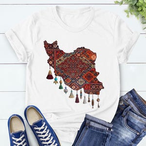 May include: White t-shirt featuring a detailed graphic of the Iran map filled with a traditional rug design in red, blue, and gold. The design includes decorative tassels. Blue sneakers and jeans are also visible.