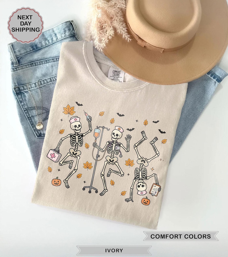 Spooky Halloween Nurse Skeleton shirt featuring dancing skeletons wearing nurse hats with medical supplies, IV drip, pumpkins, and fall leaves on an ivory tee.