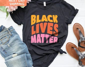 May include: Black Lives Matter t-shirt with the words in a retro font and orange, red, and pink colors. The shirt is black and is shown on a white surface.