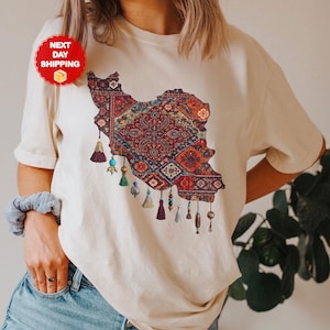 May include: Cream-colored t-shirt featuring a detailed map of Iran, filled with a colorful, intricate rug design. The design includes tassels and beads. The shirt is made of a soft, natural fabric.