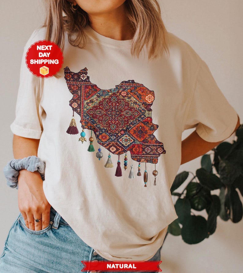 May include: Cream-colored t-shirt featuring a detailed map of Iran, filled with a colorful, intricate rug design. The design includes tassels and beads. The shirt is made of a soft, natural fabric.