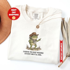 May include: Ivory-colored t-shirt featuring a cartoon frog wearing a cowboy hat and holding a coffee cup. The frog is pointing with its right hand. The text "I HAVE TO SAY WEIRD STUFF OR I'LL DIE" is printed below the frog. The shirt has a crew neck.