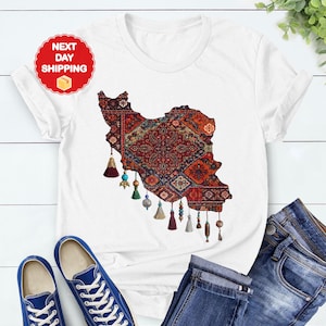 May include: White t-shirt featuring a detailed map of Iran, filled with a traditional rug design in red, blue, and gold. The shirt has decorative tassels and is paired with blue jeans and sneakers. The text "NEXT DAY SHIPPING" is visible.