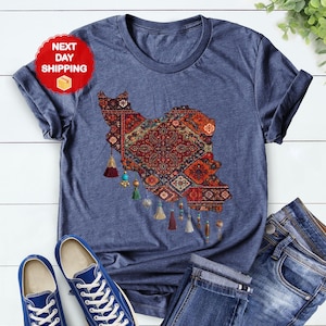 May include: Heather navy t-shirt featuring a detailed map of Iran in a traditional rug design. The map is in shades of red, orange, and blue, with decorative tassels. The shirt is paired with blue jeans and sneakers.