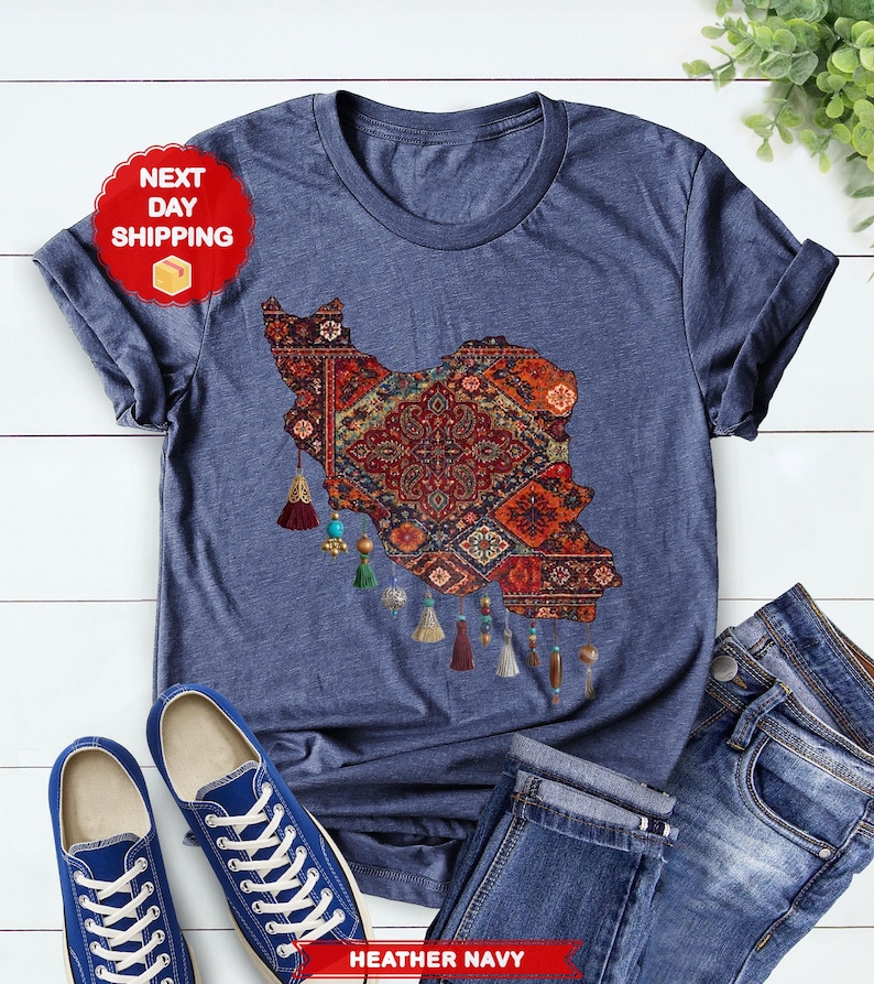 May include: Heather navy t-shirt featuring a detailed map of Iran in a traditional rug design. The map is in shades of red, orange, and blue, with decorative tassels. The shirt is paired with blue jeans and sneakers.