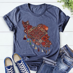 May include: Heather navy t-shirt featuring a detailed map of Iran, designed with a traditional rug pattern in shades of red, orange, and blue. The shirt includes decorative tassels and is paired with blue jeans and sneakers.