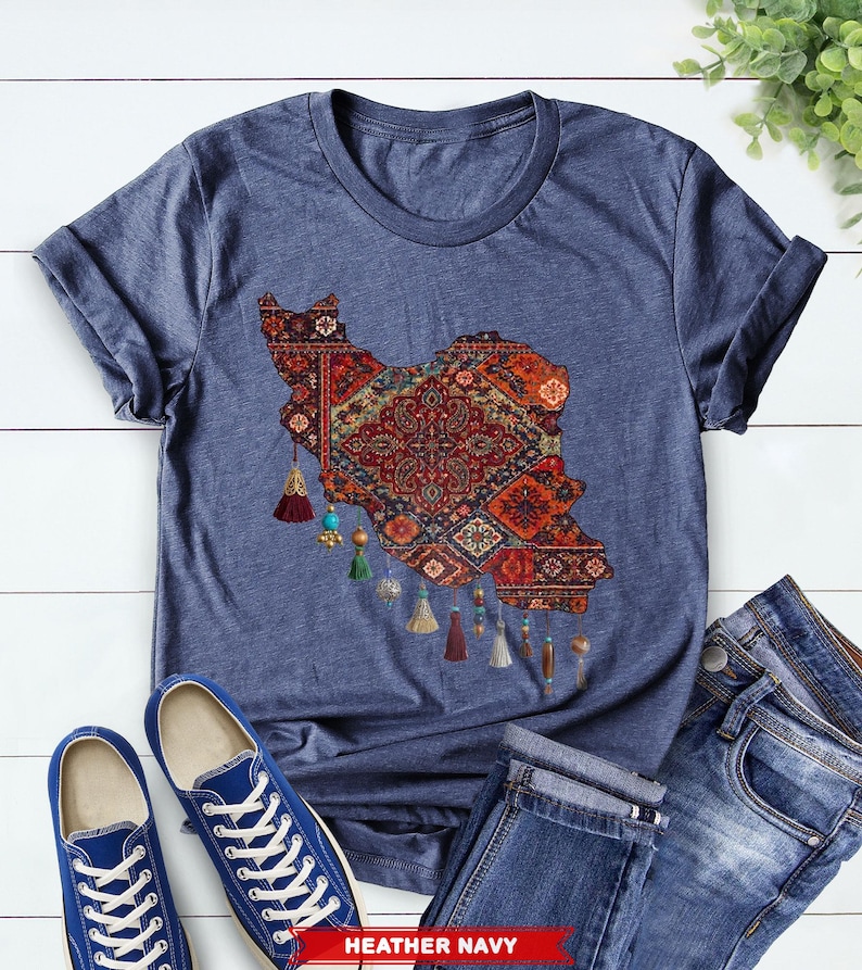 May include: Heather navy t-shirt featuring a detailed map of Iran, designed with a traditional rug pattern in shades of red, orange, and blue. The shirt includes decorative tassels and is paired with blue jeans and sneakers.