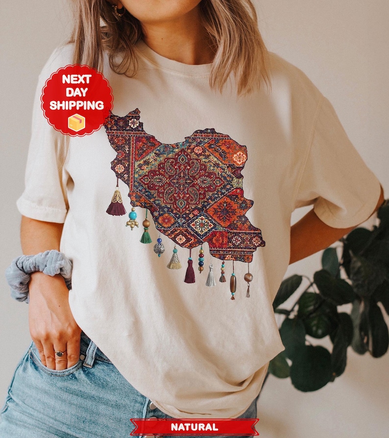 May include: Cream-colored t-shirt featuring a detailed map of Iran filled with a colorful, patterned design. The map is adorned with decorative tassels and beads. The shirt has the word "NATURAL" printed on the bottom.
