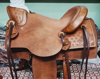 May include: A brown leather saddle with intricate floral tooling. The saddle has a high cantle and a padded seat. The saddle is on a checkered brown and white surface.